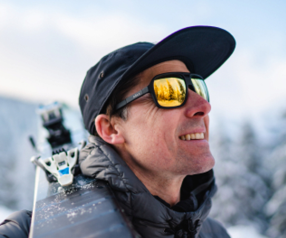 Man in snow carrying skis wearing Torino sunglasses