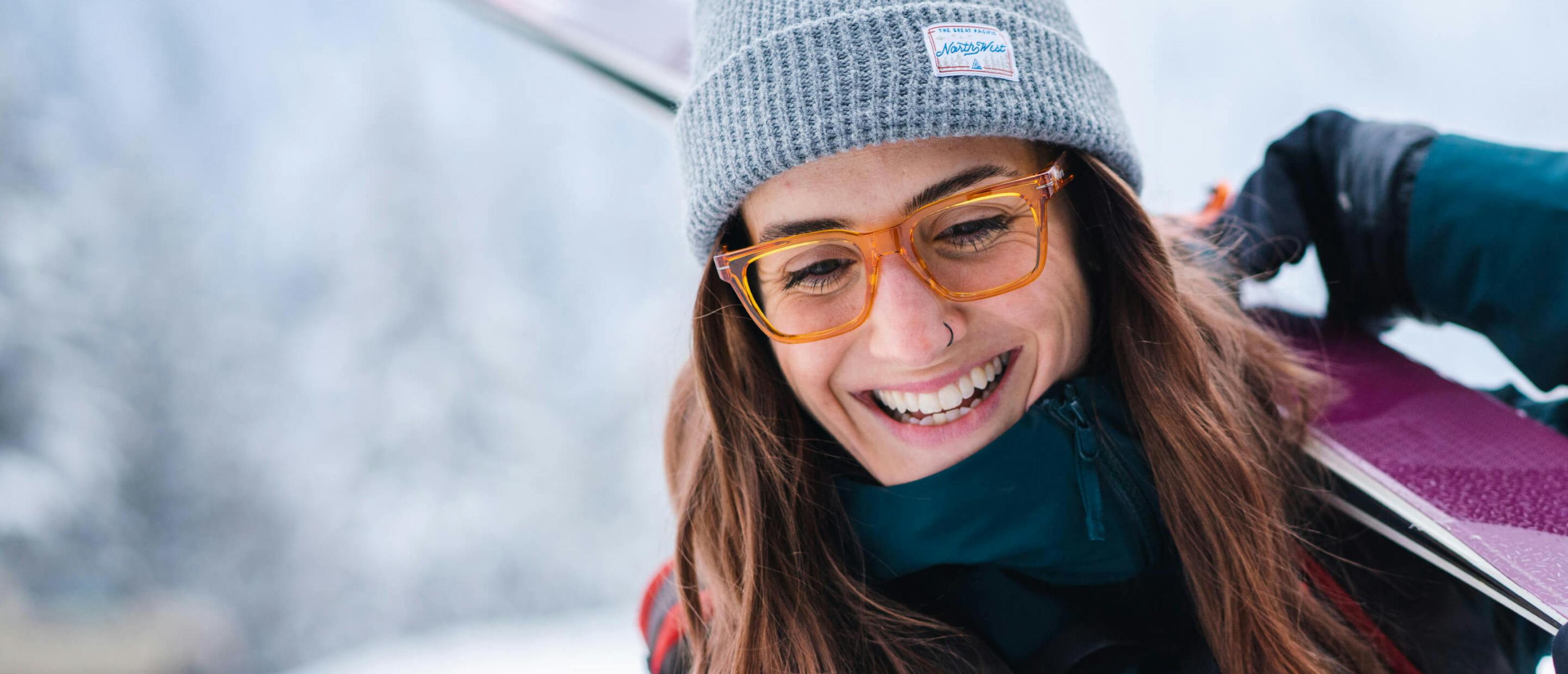 Woman smiling carrying skis wearing Lockhart orange eyeglasses