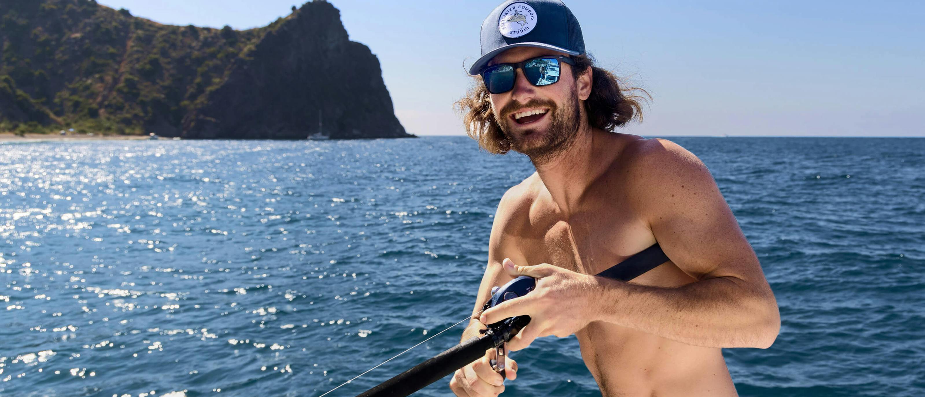 A man fishing wearing thick frames, square, matte black with glacier mirrored sunglasses in the ocean right off the coast of an island.